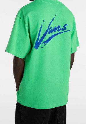 Person, der bærer en knaldgrøn T-shirt med løstsiddende ærmer og stor blå "Vans" tekst på ryggen, kombineret med sorte bukser.