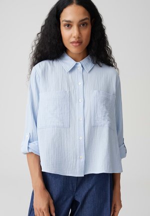 Femme aux longs cheveux bouclés portant une chemise bleu clair à boutons avec deux poches avant et manches retroussées, associée à un pantalon bleu foncé.
