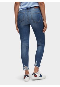 Blå smala jeans med fransad nederkant, fem fickor och en slät textur. Bärs med vita sneakers som har fjärilsapplikationer.