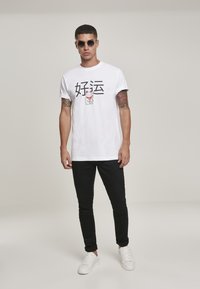 Hvit T-skjorte med svart tekst og en katt-grafikk, kombinert med svarte bukser og hvite sneakers; modellen bruker runde solbriller.