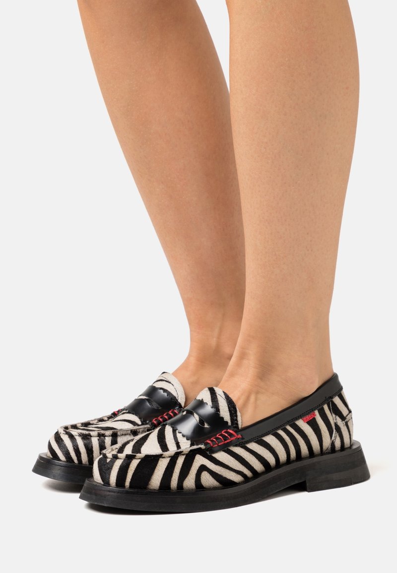 Jambes portant des mocassins rayés zèbre noir et blanc avec une bordure noire et des coutures rouges, se tenant sur un fond clair.