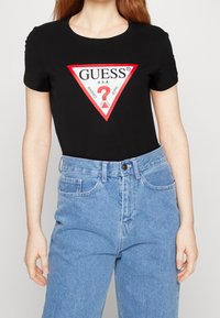 Černé bavlněné tričko s trojúhelníkovým červenobílým logem, které obsahuje slovo "GUESS" a grafické prvky. K tomu jsou světlé modré džíny.
