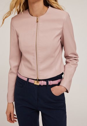 Femme portant une veste zippée rose clair, une ceinture rose assortie avec une boucle dorée, et un pantalon bleu marine foncé, main dans la poche.