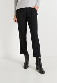 Pantalones negros ajustados con pliegues frontales, un corte relajado y bolsillos laterales, combinados con botas de tobillo negras de tacón en un fondo sencillo.