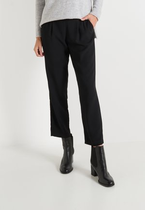 Pantalones negros ajustados con pliegues frontales, un corte relajado y bolsillos laterales, combinados con botas de tobillo negras de tacón en un fondo sencillo.