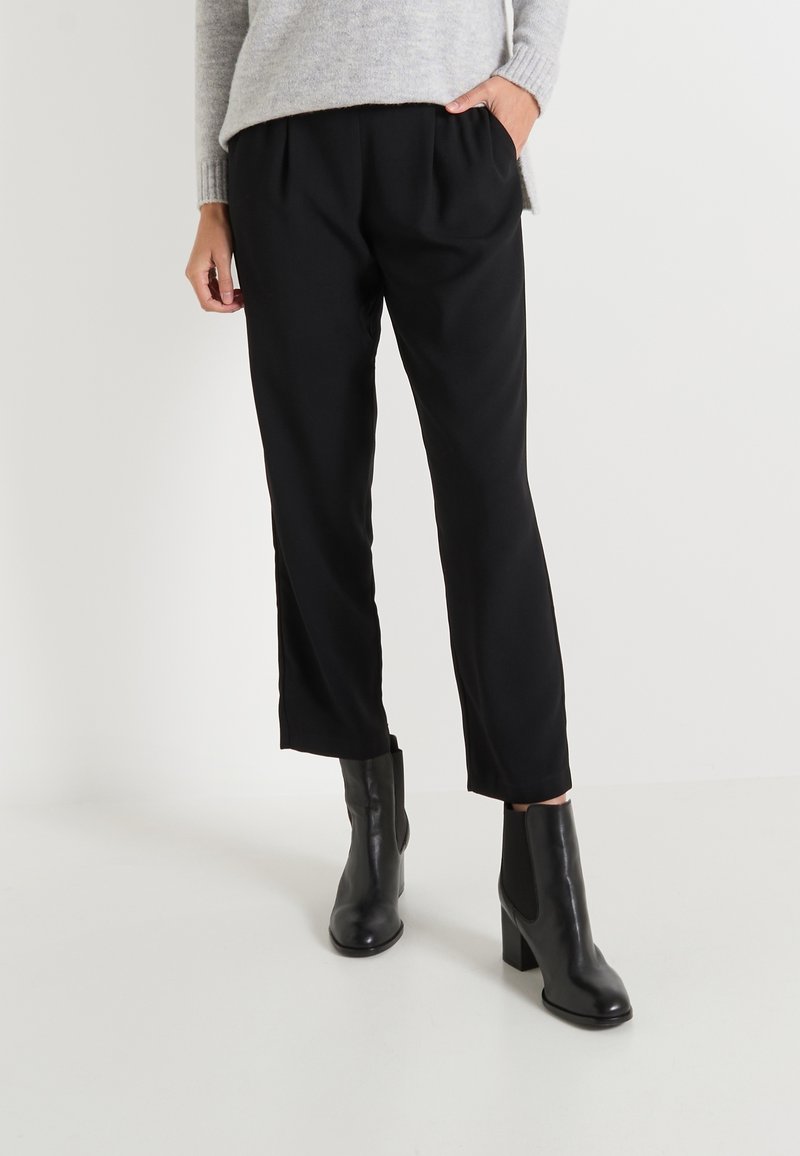 Pantalones negros ajustados con pliegues frontales, un corte relajado y bolsillos laterales, combinados con botas de tobillo negras de tacón en un fondo sencillo.