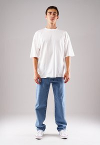 Weißes, übergroßes T-Shirt mit kurzen Ärmeln, kombiniert mit hellblauen Jeans. Die Jeans haben einen geraden Schnitt und sitzen oberhalb der Schuhe.