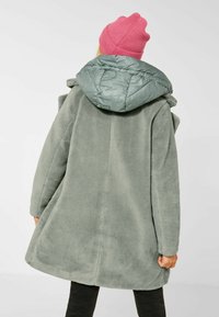 Faux fur-kappa i dämpad grön med en fluffig grå huva. Designen kännetecknas av en avslappnad passform och texturerad yta, perfekt för värme.