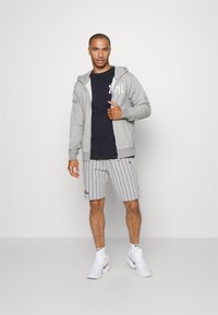 Sudadera gris con cremallera, camiseta negra, pantalones cortos de rayas grises y zapatillas blancas. La sudadera presenta un logo en el lado izquierdo del pecho.