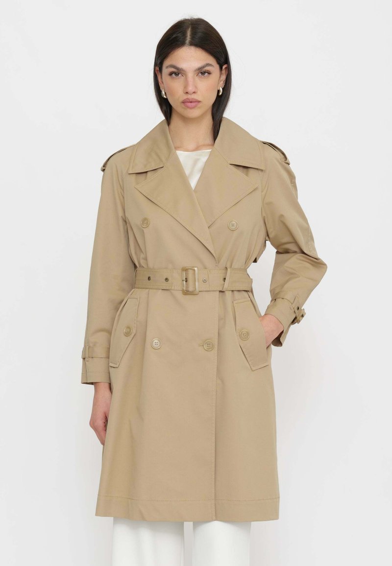 Donna che indossa un trench beige con cintura, ampi rever e tasche con bottoni, in piedi con una mano in tasca su sfondo bianco.
