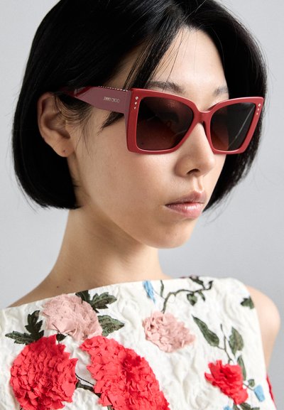 Lunettes de soleil carrées rouges avec des verres sombres, dotées d'accents en gemmes sur les branches. Le modèle porte un haut brodé de fleurs avec des couleurs vives.