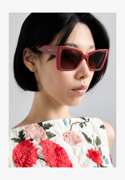 Lunettes de soleil carrées rouges avec des verres sombres, dotées d'accents en gemmes sur les branches. Le modèle porte un haut brodé de fleurs avec des couleurs vives.