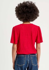 Haut rouge à manches courtes avec un motif à œillets et un tissu texturé. Encolure ronde, manches légèrement bouffantes et ourlet droit. Porté avec un jean bleu.
