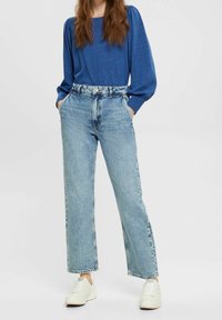 Žena ve středně světlých vysokých rovně střižených džínách, modrém dlouhém rukávu a bílých teniskách, se stojícími rukama v kapsách.