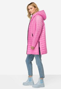 Chaqueta de plumas acolchada rosa con capucha, cierre frontal con cremallera y bolsillos laterales, combinada con botas de tobillo azul claro y vaqueros.