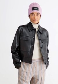 Giacca in pelle nera corta con colletto, dotata di due tasche frontali e chiusura a bottoni, indossata sopra un maglione a collo alto color crema e pantaloni a righe.