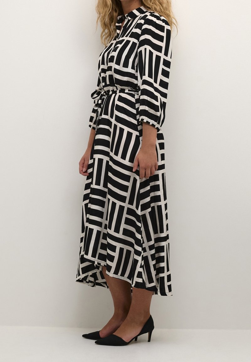Robe à motifs noir et blanc avec des manches longues, taille ceinturée et ourlet asymétrique. Portée avec des escarpins noirs à bout pointu.
