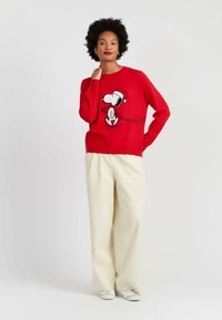 Rode gebreide trui met een Snoopy-afbeelding in zwart-wit, gecombineerd met beige wijde broekspijpen en witte sneakers.