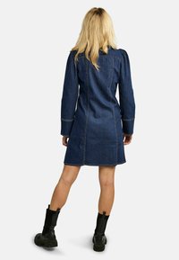 Traper haljina ima uski kroj s dugim rukavima i dužinom do sredine bedra. Tamnoplava boja s kontrastnim bijelim šavovima. Uparena s crnim čizmama.