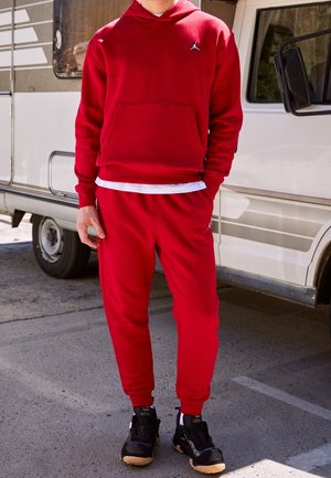 Sweat-shirt rouge à capuche avec une poche avant et un logo noir, associé à un pantalon de survêtement rouge assorti. Des chaussures de sport noires complètent l'ensemble.