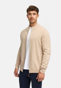 Beige rits cardigan met een ribgebreide kraag en manchetten, met een gladde textuur en minimalistisch ontwerp, gedragen over een wit shirt.