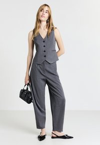 Femme portant un gilet et un pantalon à rayures gris, tenant un petit sac à main noir, se tenant avec une main derrière le dos.