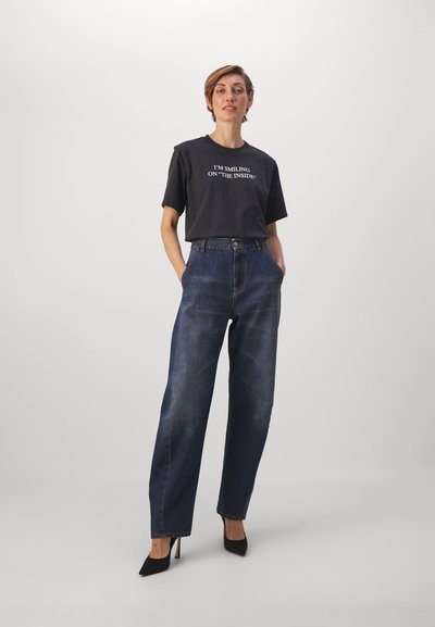 Must t-särk musta värvi valge teksti ja kõrge vöökohaga tumesiniste denim pükstega, mis on kombineeritud mustade kõrgete kontsadega. Mudel seisab käed taskutes.