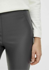 Hochtaillierte, graue Kunstlederhosen mit glatter Textur; verfügen über Seitennähte und ein enges Design; kombiniert mit einem weißen Langarmoberteil.