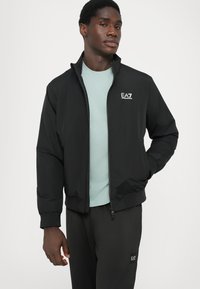 Black bomber jacket with a high collar, elastic cuffs, and a zip closure, featuring a small EA7 logo on the chest. Worn over a light blue shirt.