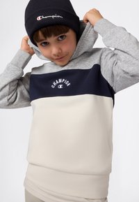Niño con sudadera de bloques de color y gorro negro ajustando su sudadera, mirando hacia la cámara sobre un fondo blanco liso.