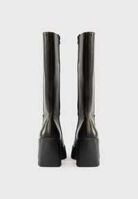 Μαύρες δερμάτινες μπότες μέχρι το γόνατο με ομαλή υφή, χοντρές σόλες πλατφόρμας και λεπτομέρειες με φερμουάρ στο πίσω μέρος. Απλός, κομψός σχεδιασμός με κατακόρυφο σχήμα.