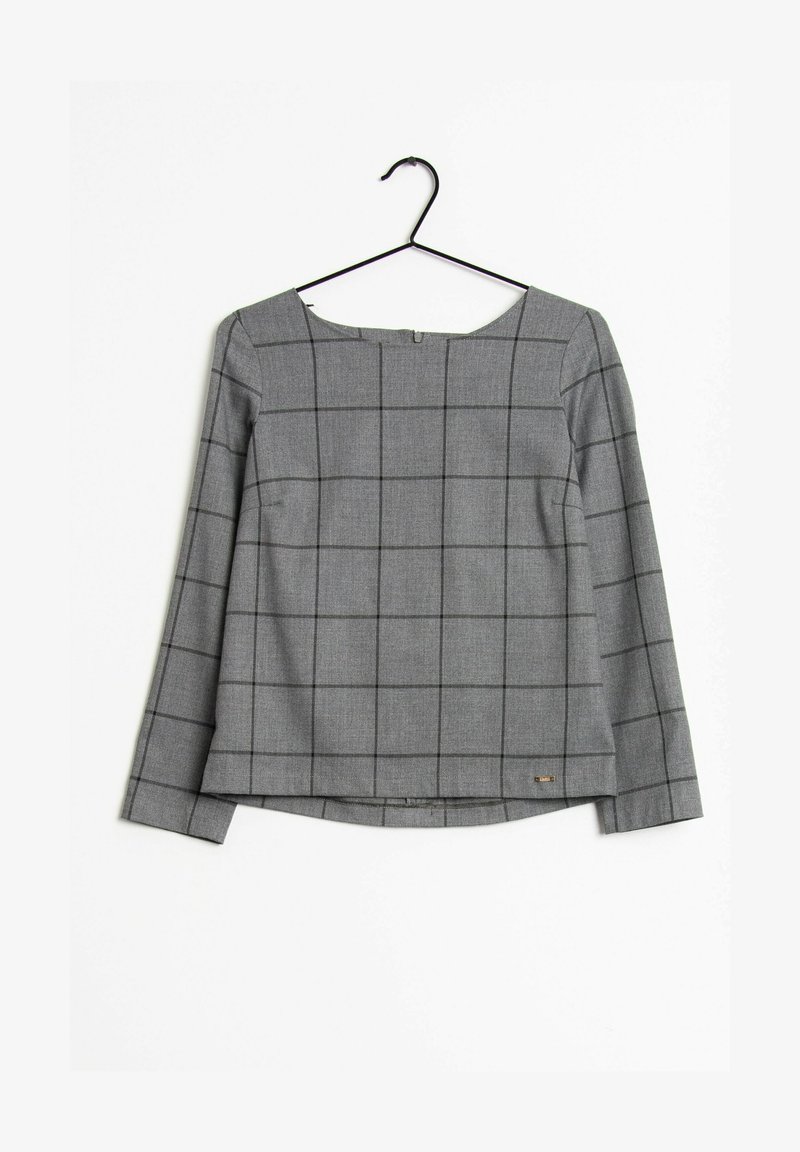 Blouse grise à manches longues avec motif à carreaux noirs, col bateau et petite étiquette métallique avec logo sur l'ourlet, suspendue à un cintre noir.