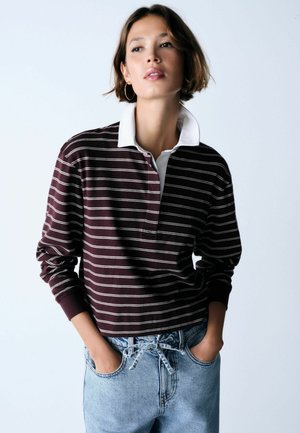 Junge Frau mit kurzen Haaren, die ein bordeauxrotes und weiß gestreiftes Rugbyshirt sowie hellblaue Jeans trägt und die Hände in den Taschen hat.