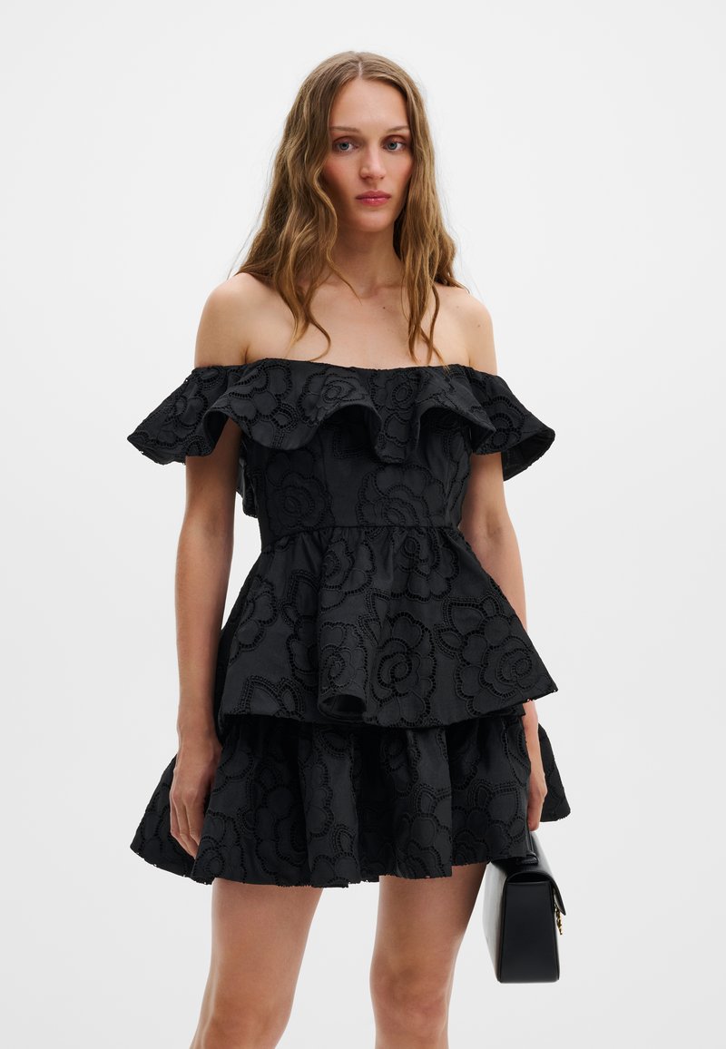 Γυναίκα με μακριά κυματιστά μαλλιά φοράει μαύρο φόρεμα με ανάγλυφα λουλούδια και ανοιχτούς ώμους και κρατά μαύρη τσάντα, σε απλό φόντο.