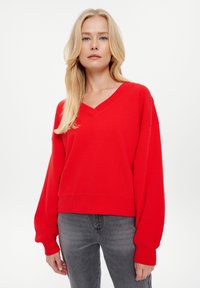 Maglione rosso a V in tessuto morbido con spalle basse e maniche lunghe, abbinato a jeans grigi. Design semplice e casual.