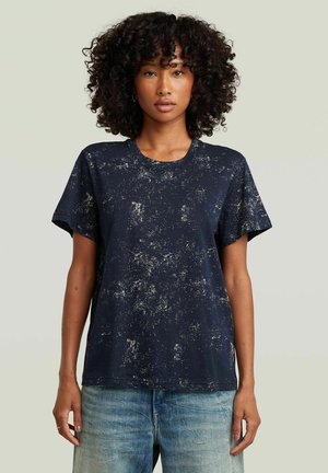 Kobieta z kręconymi włosami, ubrana w czarny T-shirt w kropki z krótkim rękawem i niebieskie dżinsy, stojąca z ramionami swobodnie opuszczonymi wzdłuż ciała.