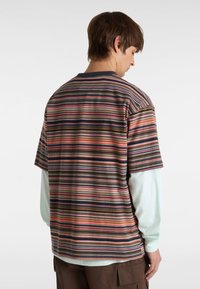 Camisa de manga corta a rayas en marrón, naranja, azul marino y rosa. Cuenta con cuello redondo y un corte relajado, llevada sobre una camiseta de manga larga de color azul claro.