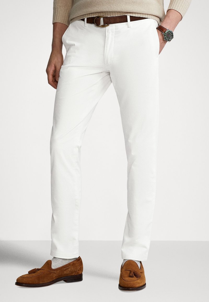Homme portant un pantalon blanc, une ceinture beige, un pull beige, des mocassins à pampilles en daim marron, des chaussettes grises et une montre-bracelet avec un bracelet en cuir marron.