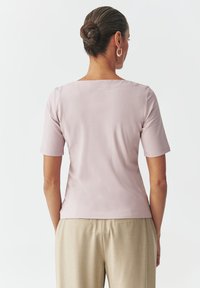 Camicia a maniche corte di colore rosa chiaro, con scollo a quadrato e texture liscia, abbinata a pantaloni beige per un look coordinato.