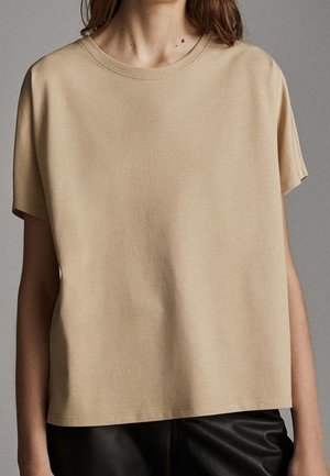 Femme portant un t-shirt beige uni à manches courtes et col rond, associé à un pantalon en cuir noir sur un fond neutre.
