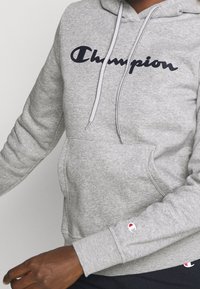 Mörkhyad person iklädd en grå Champion-hoodie med framficka, logotyp på bröstet och ärmen, samt svarta byxor med logotyp delvis synlig.