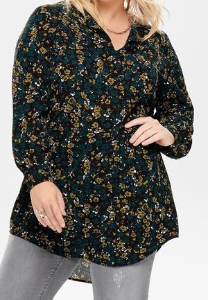 Nainen, jolla on päällään pitkähihaisen mustan paidan vihreällä, keltaisella ja valkoisella kukkakuvioinnilla, yhdistettynä vaaleanharmaisiin kuluneisiin farkkuihin.