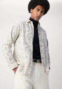 Camisa de rayas paisley de color beige con mangas largas, cierre de botones y cuello. Llevada sobre un suéter de cuello alto negro. Combinada con pantalones de un color claro.