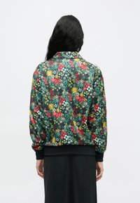 Person mit langen schwarzen Haaren, der eine bunte Blumjacke mit roten, gelben, grünen und weißen Blumen über einem schwarzen Rock trägt, von hinten gesehen.