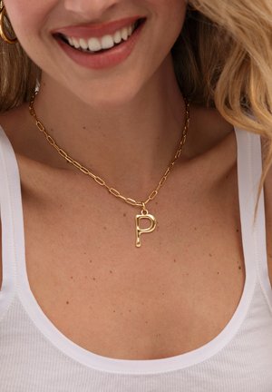 Mujer sonriente con camiseta sin mangas blanca y collar de cadena dorada con un colgante grande en forma de letra "P".