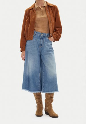 Person wears brown suede jacket, tan knit polo, wide-leg faded blue jeans, and tan suede mid-calf boots with decorative studs.