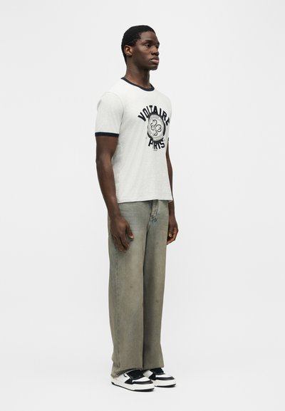 Νεαρός άνδρας όρθιος, φοράει λευκό T-shirt με την επιγραφή "Voltaire Paris" και μαύρη περιμέτρου, γκρι φαρδιά τζιν και ασπρόμαυρα αθλητικά παπούτσια, μπροστά σε απλό φόντο.