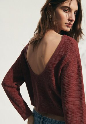 Giovane donna con capelli castani che indossa un maglione rosso scuro con trama e scollatura sulla schiena, posando con una mano sui fianchi davanti a uno sfondo neutro.