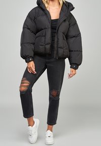 Chaqueta negra acolchada con un diseño corto y capucha, combinada con jeans de mezclilla negra desgastados con rodilleras rasgadas y zapatillas blancas.