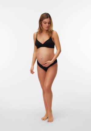 Meztelen lábú terhes nő áll, fekete fehérneműt visel, finoman átöleli a hasát, lefelé néz, tiszta fehér háttér előtt.